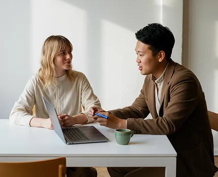 Two colleagues discussing work at a table with a laptop and coffee cup.