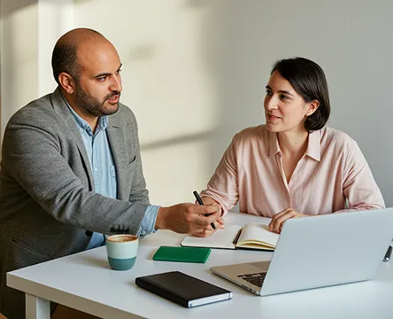 Man and woman discussing notes at a desk with a laptop and notebooks.